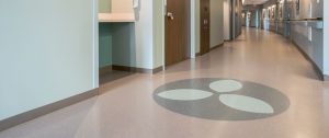 Seamless commercial resilient sheet flooring with a custom circular leaf logo inlay and integrated baseboards by All Floors Commercial.